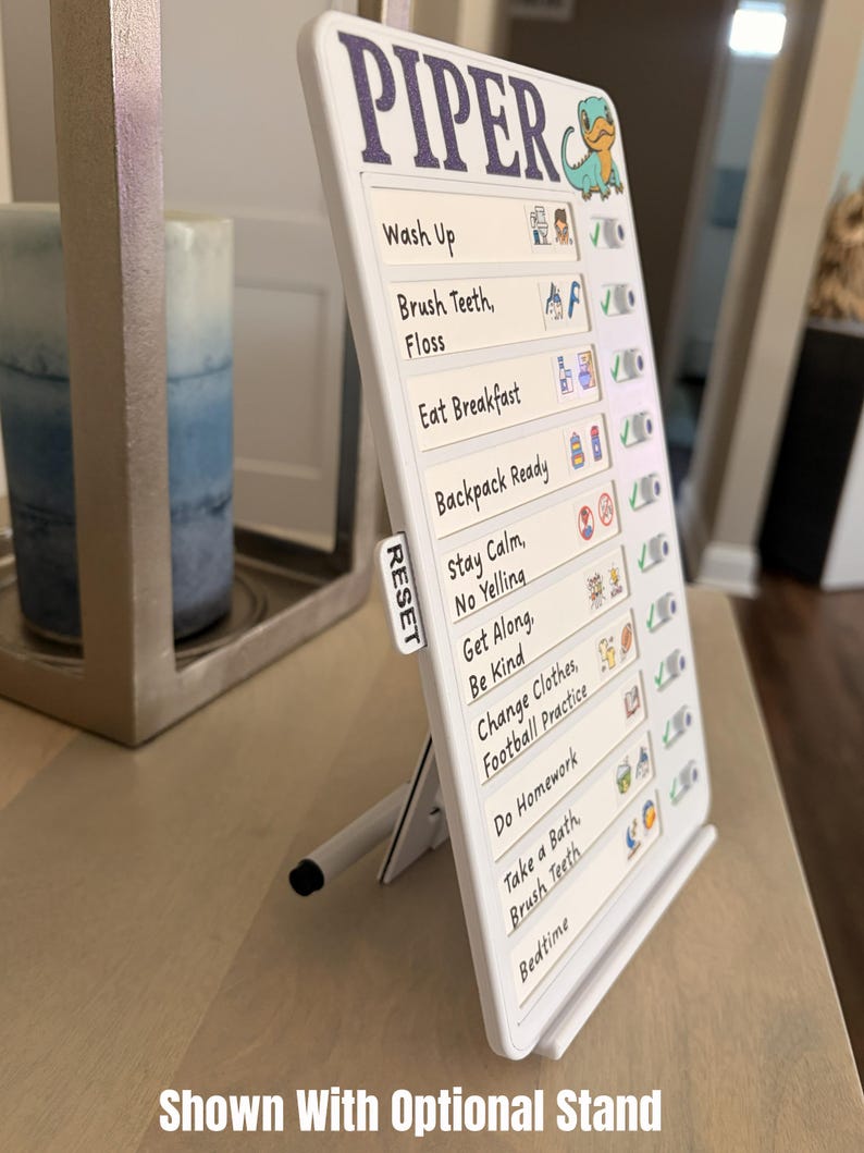 May include: White chore chart with the name "PIPER" in purple. The chart includes a list of tasks with check boxes and a small dinosaur graphic. The chart is propped up by a white stand. The text "Shown With Optional Stand" is at the bottom.