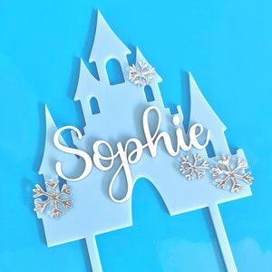 May include: A pale blue castle cake topper with the name "Sophie" in white script. Silver snowflake embellishments are placed around the castle design. The topper has two sticks for insertion into a cake.
