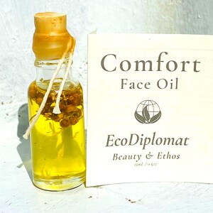 May include: A small glass bottle with a cork stopper filled with yellow oil and dried flowers. The bottle is labeled "Comfort Face Oil" and "EcoDiplomat Beauty & Ethos 15ml / 0.5oz"