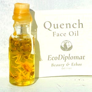 May include: A small glass bottle with a yellow cork and a white string tied around the neck. The bottle is filled with a yellow liquid and dried flowers. The bottle is labeled "Quench Face Oil" and "EcoDiplomat Beauty & Ethos 15ml / 0.5oz".