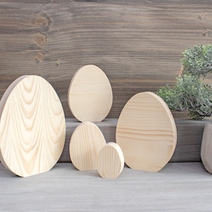 Może przedstawiać: Pięć drewnianych ozdób w kształcie jajka w różnych rozmiarach, ułożonych na jasnoszarym tle. Największa ozdoba znajduje się po lewej stronie, a najmniejsza po prawej. Ozdoby są wykonane z naturalnego drewna i mają jasnobrązowy kolor.