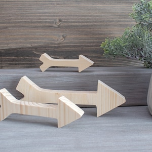 Könnte beinhalten: Vier unfertige Holzpfeile in verschiedenen Größen. Die Pfeile sind aus hellfarbigem Holz gefertigt und haben ein schlichtes, klares Design. Die Pfeile sind auf einer grauen Oberfläche mit einem Holz Hintergrund und einer kleinen Pflanze in einem Topf angeordnet.