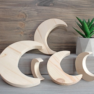 Può includere: Quattro forme di luna crescente in legno grezzo di diverse dimensioni. Le lune sono disposte su una superficie grigio chiaro. Una piccola pianta grassa in vaso si trova sullo sfondo.