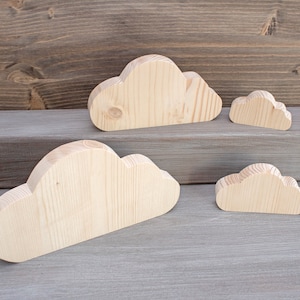 Puede incluir: Cuatro formas de nubes de madera de diferentes tamaños, dispuestas sobre una superficie de madera gris. Las nubes son de madera natural y tienen un color marrón claro.