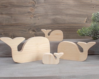 May include: Four wooden whale ornaments in various sizes. The largest whale is light brown and has a simple, rounded shape. The other whales are smaller and have more detailed features.