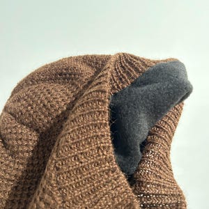 K&ouml;nnte beinhalten: Eine braune Strickm&uuml;tze mit einem dunkelgrauen Fleecefutter. Die M&uuml;tze besteht aus einem strukturierten Material mit offener Webart. Das Innenfutter ist sichtbar und bietet W&auml;rme und Komfort.