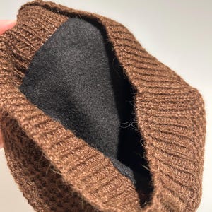 K&ouml;nnte beinhalten: Eine braune Strickm&uuml;tze mit schwarzem Innenfutter. Die M&uuml;tze besteht aus einem strukturierten, gerippten Strickmaterial. Das Innenfutter ist dunkel und einfarbig. Die M&uuml;tze scheint umgeschlagen zu sein.