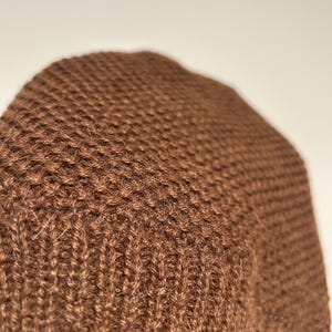 K&ouml;nnte beinhalten: Nahaufnahme einer braunen Strickm&uuml;tze. Die M&uuml;tze besteht aus einem strukturierten, gewebten Material. Die Farbe ist ein sattes, warmes Braun, und das Strickmuster ist im gesamten Stoff sichtbar.