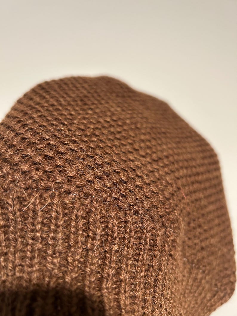 K&ouml;nnte beinhalten: Nahaufnahme einer braunen Strickm&uuml;tze. Die M&uuml;tze besteht aus einem strukturierten, gewebten Material. Die Farbe ist ein sattes, warmes Braun, und das Strickmuster ist im gesamten Stoff sichtbar.