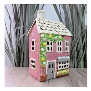 May include: A pink ceramic house with a white roof, chimney, and window frames. The house has a grey door and the text "FLOWER SHOP" above the windows. Decorative flowers and vines adorn the facade.