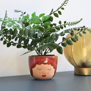 May include: A brown ceramic planter shaped like a woman's face with closed eyes, red lips, and a red and white polka dot headband. The planter is filled with a green leafy plant.