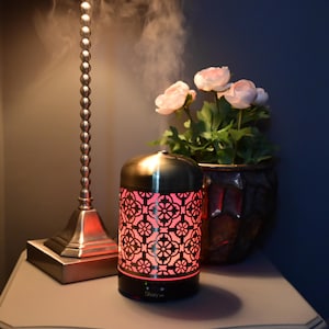 May include: A gold metal essential oil diffuser with a red light and a patterned design. The diffuser is on a table with a lamp and flowers.