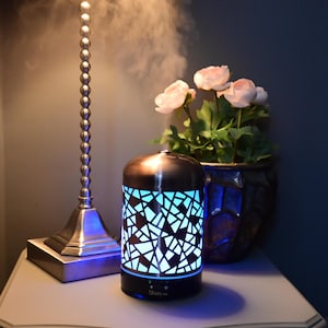 May include: A stainless steel essential oil diffuser with a geometric pattern and a blue light. The diffuser is on a white bedside table with a lamp and flowers.