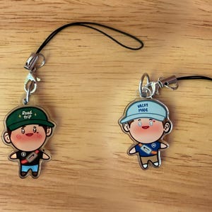 May include: Two acrylic character charms with black phone straps. The left charm features a figure in a green cap with the text Road trip. The right charm features a figure in a light blue cap with the text VACAY MODE. Both figures have rosy cheeks.