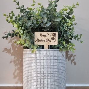 May include: A white planter with a textured surface holds a green potted plant with a wooden sign that reads "Happy Mothers Day" with a dandelion design.