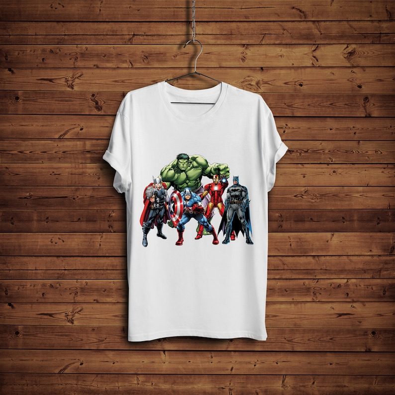 May include: White t-shirt featuring a graphic of several superheroes, including the Hulk, Thor, Captain America, Iron Man, and Batman. The shirt is hanging on a hanger against a wooden background. The superheroes are in their iconic costumes.