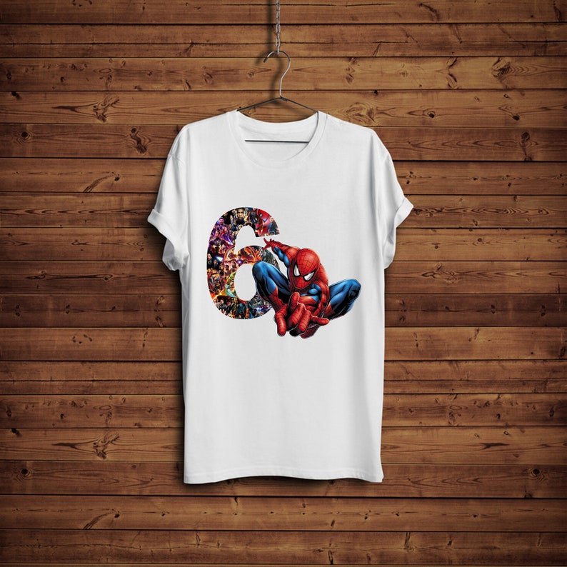 Op de afbeelding: Wit t-shirt met een afbeelding van Spider-Man in een hurkende pose. Het nummer "6" is op het shirt gedrukt, gevuld met verschillende stripfiguren. Het t-shirt hangt aan een metalen hanger tegen een houten achtergrond.