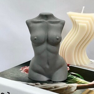 Puede incluir: Una vela gris en forma de torso con un acabado liso y mate. La vela está sentada sobre un libro con un diseño floral.