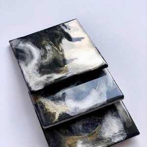 Puede incluir: Tres posavasos cuadrados con un diseño abstracto en negro, blanco y dorado. Los posavasos están apilados uno encima del otro.