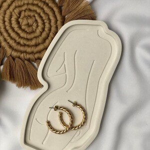 Puede incluir: Una bandeja de cerámica blanca con forma de figura humana con dos aros de oro en la parte superior.