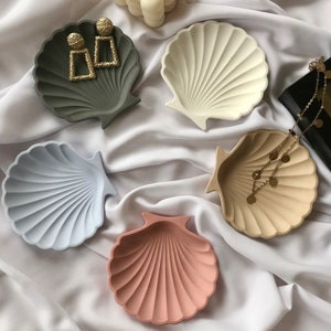 Könnte beinhalten: Eine Sammlung muschelförmiger Tabletts in verschiedenen Farben wie Grün, Weiß, Blau, Rosa und Beige. Einige Tabletts enthalten goldene Ohrringe und eine Halskette. Eine cremefarbene Blasenkerze ist ebenfalls vorhanden.