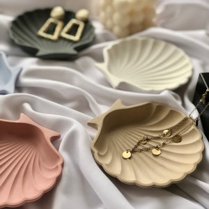 Puede incluir: Una colecci&oacute;n de cuencos en forma de concha marina en varios colores, incluyendo rosa, beige y gris. Pendientes dorados y un delicado collar dorado con dijes de disco se exhiben en los cuencos. Una vela blanca est&aacute; en el fondo.