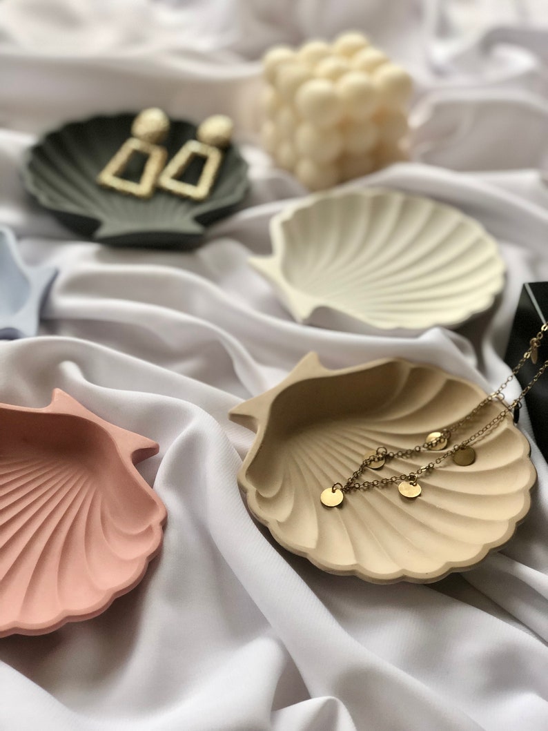 Puede incluir: Una colecci&oacute;n de cuencos en forma de concha marina en varios colores, incluyendo rosa, beige y gris. Pendientes dorados y un delicado collar dorado con dijes de disco se exhiben en los cuencos. Una vela blanca est&aacute; en el fondo.