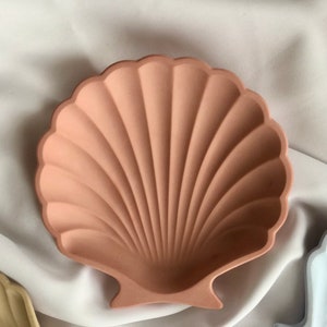 Puede incluir: Un plato decorativo en forma de concha de color melocot&oacute;n. El plato tiene un borde festoneado y un interior acanalado, que se asemeja a una concha real. El plato est&aacute; hecho de un material s&oacute;lido y probablemente se utiliza con fines decorativos o para guardar objetos peque&ntilde;os.