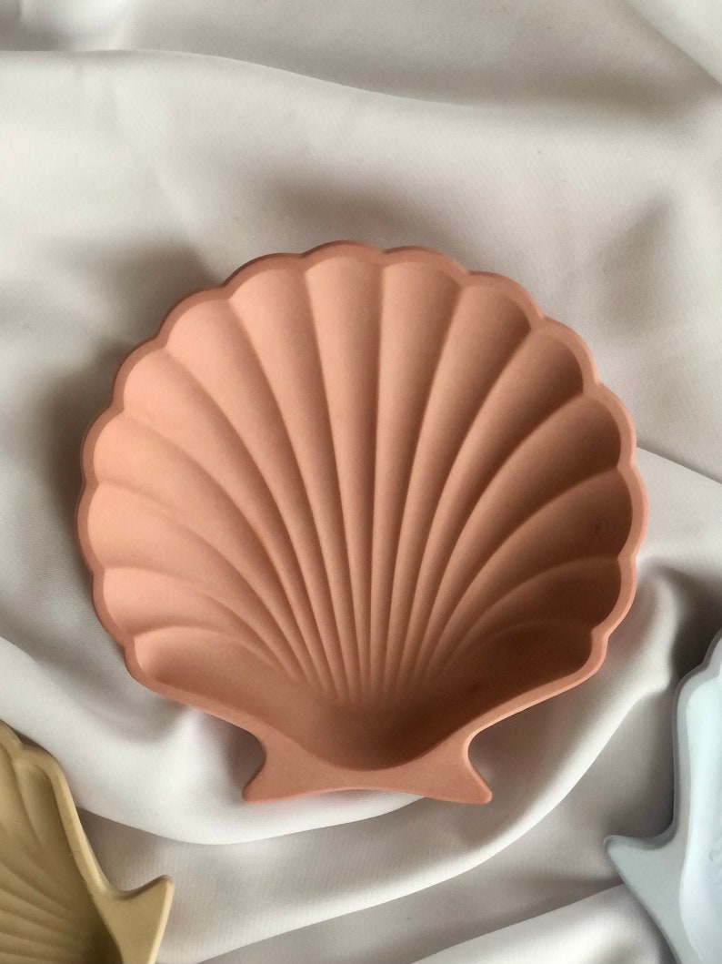 Puede incluir: Un plato decorativo en forma de concha de color melocot&oacute;n. El plato tiene un borde festoneado y un interior acanalado, que se asemeja a una concha real. El plato est&aacute; hecho de un material s&oacute;lido y probablemente se utiliza con fines decorativos o para guardar objetos peque&ntilde;os.