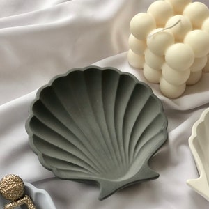 Puede incluir: Un cuenco gris en forma de concha con una superficie texturizada. Una vela blanca con forma de burbujas en el fondo. El cuenco es de hormig&oacute;n y es adecuado para guardar peque&ntilde;as joyas.
