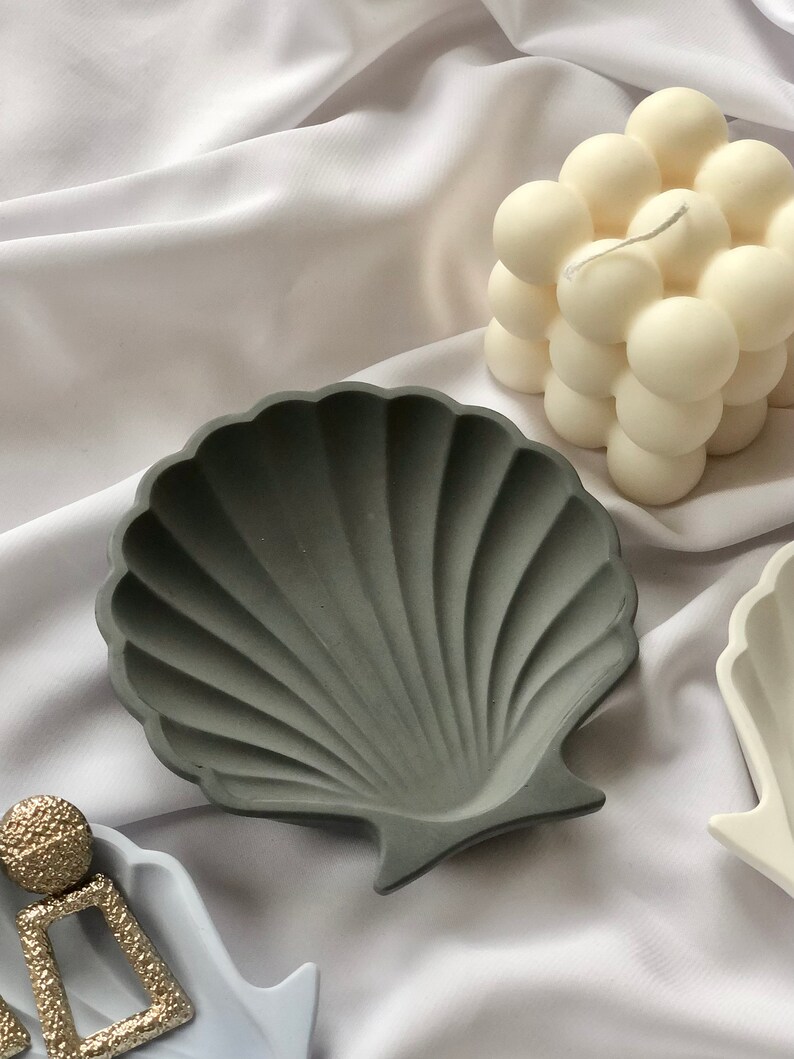 Puede incluir: Un cuenco gris en forma de concha con una superficie texturizada. Una vela blanca con forma de burbujas en el fondo. El cuenco es de hormig&oacute;n y es adecuado para guardar peque&ntilde;as joyas.