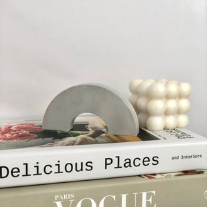 Puede incluir: Dos libros apilados uno encima del otro. El libro superior es blanco con el texto "Delicious Places and Interiors" impreso en negro. El libro inferior es beige con el texto "PARIS VOGUE" impreso en blanco. Una vela blanca de burbujas y un arco de hormigón gris están encima de los libros.
