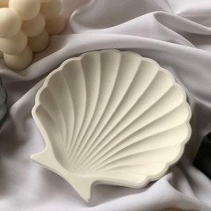 Puede incluir: Plato decorativo en forma de concha de color blanco roto con un dise&ntilde;o acanalado. El plato est&aacute; hecho de un material s&oacute;lido y se encuentra sobre una tela blanca. Es un art&iacute;culo de decoraci&oacute;n del hogar de tem&aacute;tica costera.