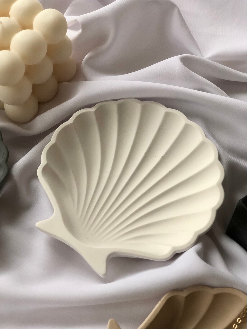 Puede incluir: Plato decorativo en forma de concha de color blanco roto con un dise&ntilde;o acanalado. El plato est&aacute; hecho de un material s&oacute;lido y se encuentra sobre una tela blanca. Es un art&iacute;culo de decoraci&oacute;n del hogar de tem&aacute;tica costera.