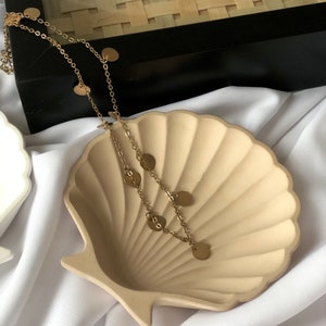 Puede incluir: Un collar dorado con peque&ntilde;os dijes redondos descansa sobre un plato beige en forma de concha. El plato est&aacute; sobre un fondo de tela blanca, con una caja negra y una textura tejida en el fondo. El collar es delicado y elegante.
