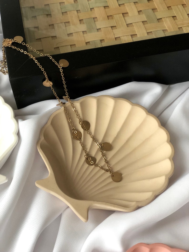 Puede incluir: Un collar dorado con peque&ntilde;os dijes redondos descansa sobre un plato beige en forma de concha. El plato est&aacute; sobre un fondo de tela blanca, con una caja negra y una textura tejida en el fondo. El collar es delicado y elegante.