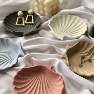 Puede incluir: Una colecci&oacute;n de cuencos en forma de concha marina en varios colores, incluyendo gris, azul claro, crema y rosa. Algunos cuencos contienen joyas, como pendientes dorados y un delicado collar. Una vela blanca con forma de burbujas est&aacute; en el fondo.