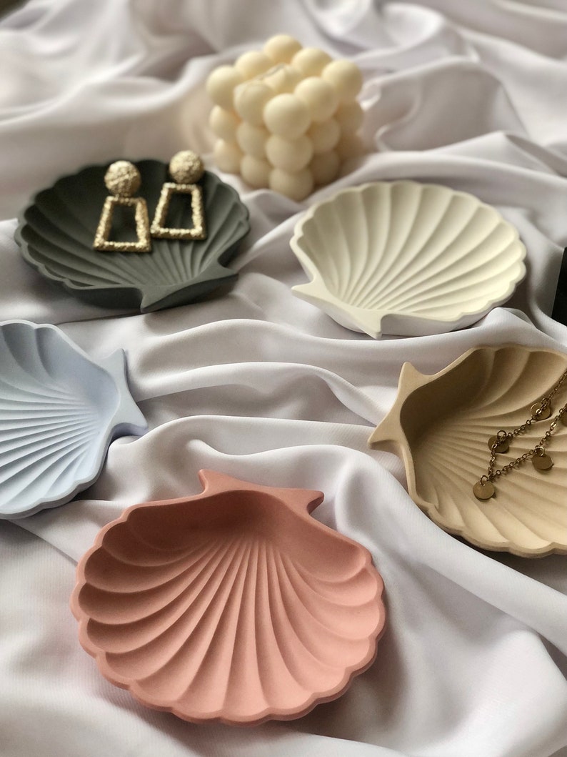 Puede incluir: Una colecci&oacute;n de cuencos en forma de concha marina en varios colores, incluyendo gris, azul claro, crema y rosa. Algunos cuencos contienen joyas, como pendientes dorados y un delicado collar. Una vela blanca con forma de burbujas est&aacute; en el fondo.