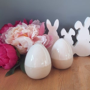 May include: Two white ceramic Easter eggs with a glossy finish, sitting on a wooden surface in front of a white ceramic bunny rabbit cutout.