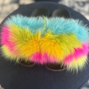 May include: A pair of fuzzy slides with a multi-colored fur design. The fur is a mix of blue, yellow, and pink.