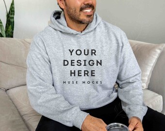 May include: A gray hooded sweatshirt with the text "YOUR DESIGN HERE MUSE MOCKS" printed on the front. The person is wearing black pants and holding a black travel mug.