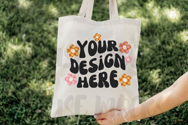 Op de afbeelding: Witte canvas draagtas met de tekst "YOUR DESIGN HERE" in zwart, retro-lettertype. De tekst wordt omringd door kleurrijke, geruite bloemen.