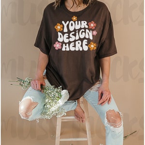 May include: A brown t-shirt with the text "YOUR DESIGN HERE" in white, with orange and pink flower graphics. The model wears light blue ripped jeans and holds a bouquet of white flowers.