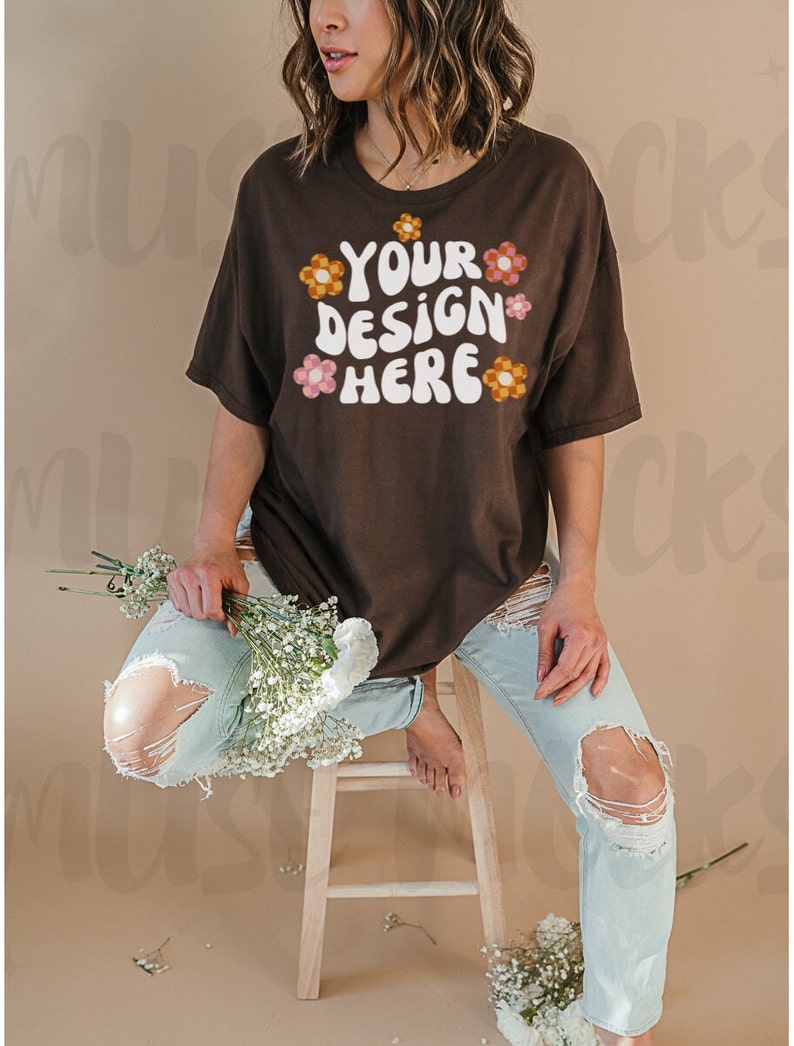May include: A brown t-shirt with the text "YOUR DESIGN HERE" in white, with orange and pink flower graphics. The model wears light blue ripped jeans and holds a bouquet of white flowers.