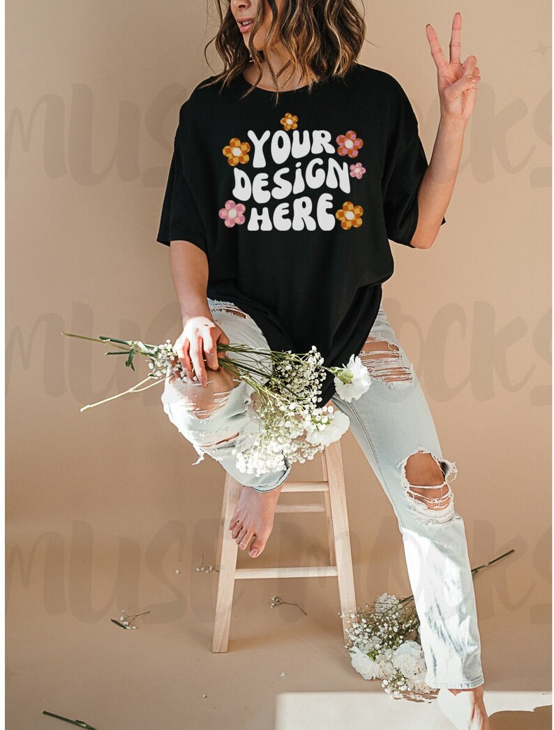 May include: Black t-shirt with the text "YOUR DESIGN HERE" in a retro font, decorated with floral accents. The model is wearing light wash ripped jeans and holding white flowers. The model is sitting on a wooden stool.