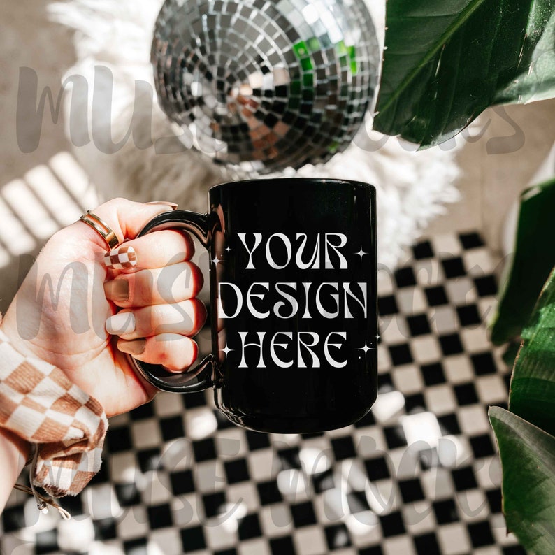 Puede incluir: Una taza de cer&aacute;mica negra con un dise&ntilde;o blanco que dice "YOUR DESIGN HERE" en una fuente retro. La taza est&aacute; siendo sostenida por una mano con una manga a cuadros.