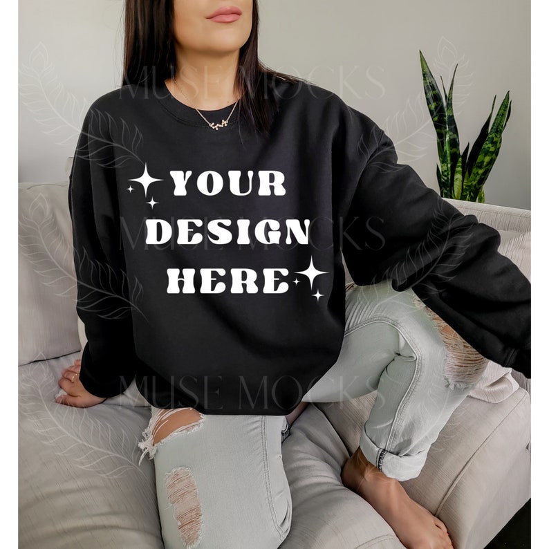 May include: A black sweatshirt with the text "YOUR DESIGN HERE" in white, with small stars on either side of the text. The sweatshirt is being worn by a person sitting on a couch.