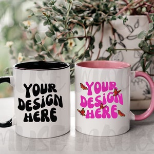 May include: Two white ceramic mugs with black and pink accents. The mug on the left has a black rim and handle and the text "YOUR DESIGN HERE" in black. The mug on the right has a pink rim and handle and the text "YOUR DESIGN HERE" in pink with small red butterflies around the text.