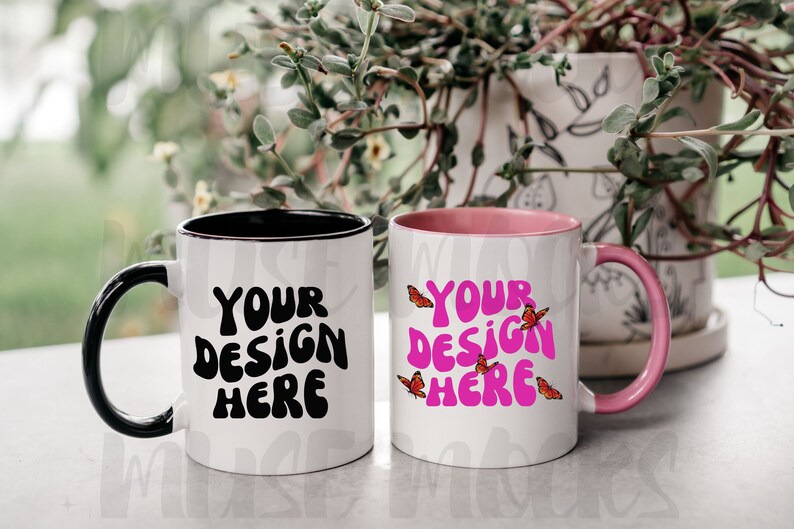 May include: Two white ceramic mugs with black and pink accents. The mug on the left has a black rim and handle and the text "YOUR DESIGN HERE" in black. The mug on the right has a pink rim and handle and the text "YOUR DESIGN HERE" in pink with small red butterflies around the text.