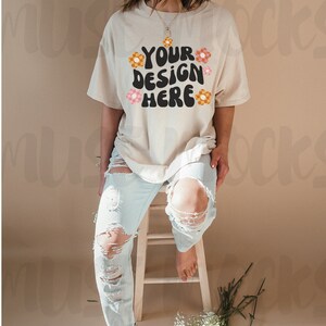 May include: A beige t-shirt with the text "YOUR DESIGN HERE" in black, decorated with orange and pink flowers. The model wears light blue ripped jeans and sits on a wooden stool. White flowers and greenery are on the floor.